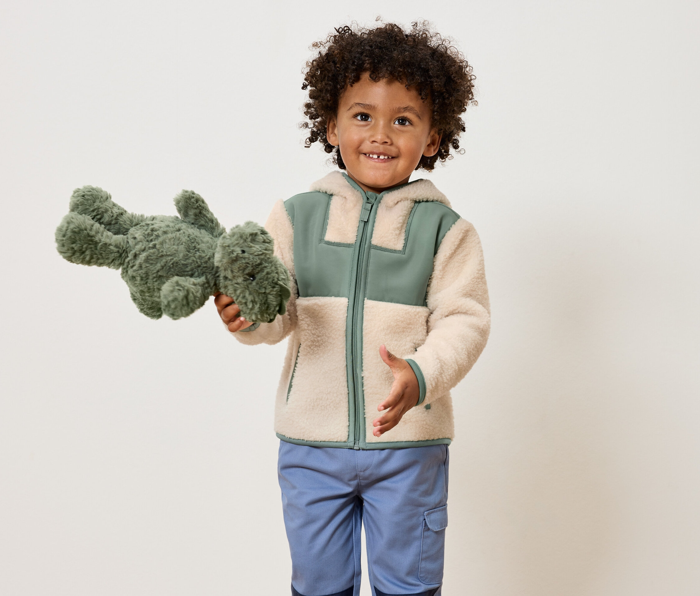 Un garçon souriant aux cheveux bouclés tient un dinosaure en peluche vert et porte une veste marron clair avec des touches de vert et un pantalon bleu.