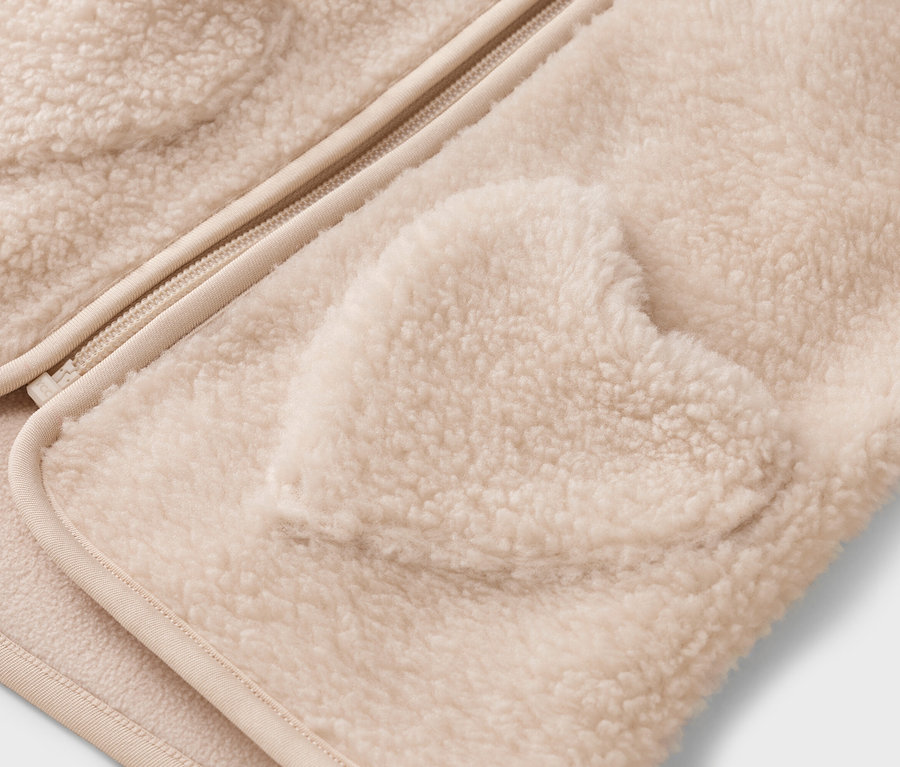 Gros plan d'une veste teddy crème avec fermeture éclair et poche en forme de cœur.