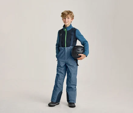 Garçon souriant portant un pantalon de ski bleu avec des bretelles noires et une veste polaire bleue et noire, tenant un casque de ski noir.