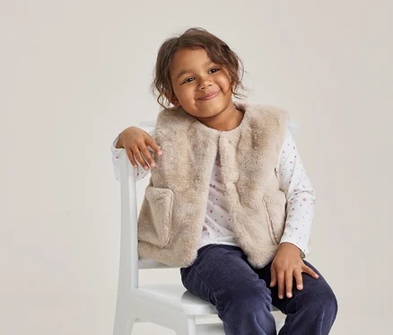 Jeune fille aux cheveux bouclés foncés souriant, portant un gilet en fausse fourrure beige clair, un haut blanc à manches longues avec des motifs roses et un pantalon bleu foncé, assise sur une chaise blanche.