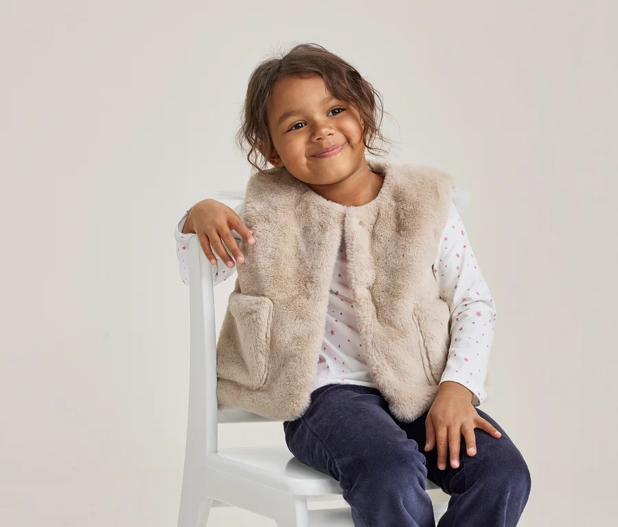 Jeune fille aux cheveux bouclés foncés souriant, portant un gilet en fausse fourrure beige clair, un haut blanc à manches longues avec des motifs roses et un pantalon bleu foncé, assise sur une chaise blanche.