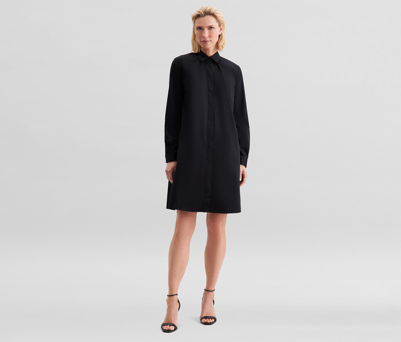 Femme blonde debout, portant une robe chemise noire à manches longues et des sandales noires à talons hauts.