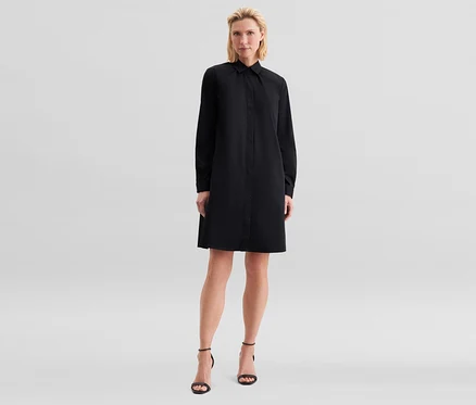 Femme blonde debout, portant une robe chemise noire à manches longues et des sandales noires à talons hauts.
