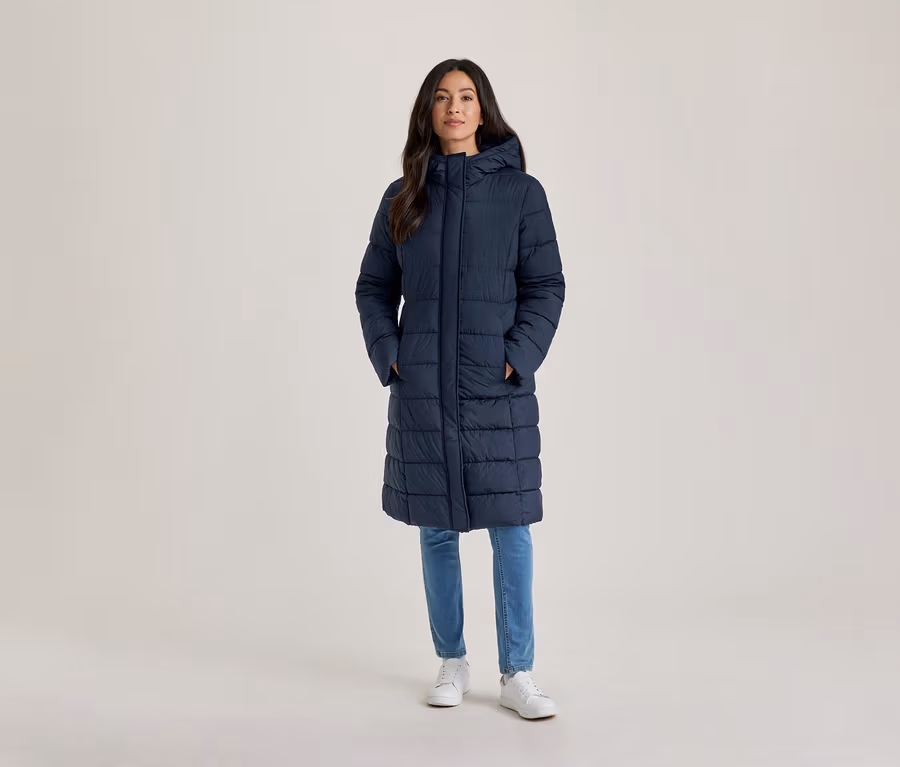 Femme debout, portant un long manteau doudoune bleu marine à capuche, un jean bleu clair et des baskets blanches, les mains dans les poches.