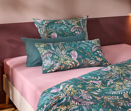 Un lit avec un drap-housse rose et une parure de lit verte à motifs floraux et de feuilles. Trois oreillers sont disposés en tête de lit, deux à motifs et un vert uni. Une table de chevet en bois avec un vase blanc et une fleur se trouve à gauche, devant un mur bicolore.