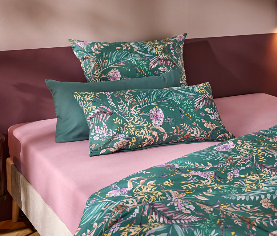 Un lit avec un drap-housse rose et une parure de lit verte à motifs floraux et de feuilles. Trois oreillers sont disposés en tête de lit, deux à motifs et un vert uni. Une table de chevet en bois avec un vase blanc et une fleur se trouve à gauche, devant un mur bicolore.