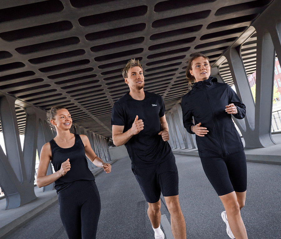 Trois personnes, deux femmes et un homme, courent sous un pont. La femme à gauche porte un top de sport noir et un legging de sport « ActivePower » noir. L'homme au milieu porte un T-shirt de sport noir et un short de sport noir. La femme à droite porte une veste coupe-vent « packable » noire et un short de sport noir.