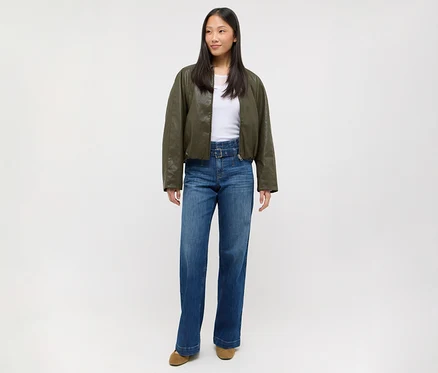 Femme debout, portant une veste bomber vert foncé, un haut blanc, un jean bleu large avec une ceinture et des chaussures marron clair.