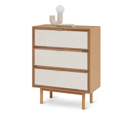 Commode en bois clair à trois tiroirs avec façades en tissu crème et poignées dorées. Une lampe sphérique moderne et des livres sont posés dessus.