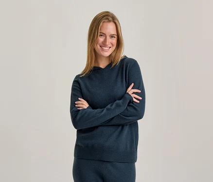 Femme souriante aux cheveux blonds portant un sweat à capuche bleu foncé et un pantalon assorti, debout les bras croisés devant un fond uni.