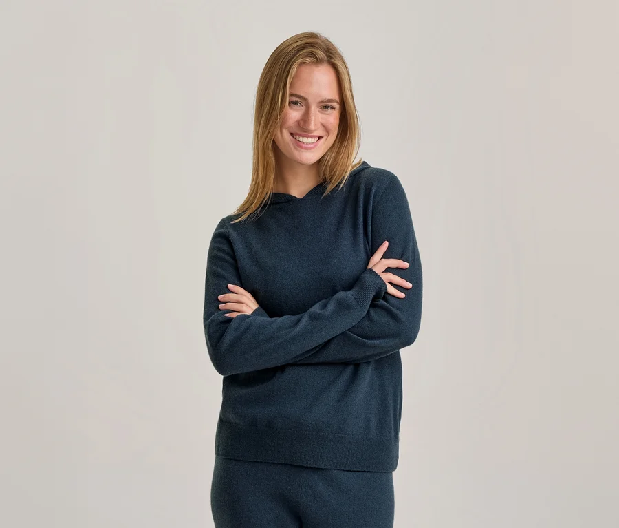 Femme souriante aux cheveux blonds portant un sweat à capuche bleu foncé et un pantalon assorti, debout les bras croisés devant un fond uni.