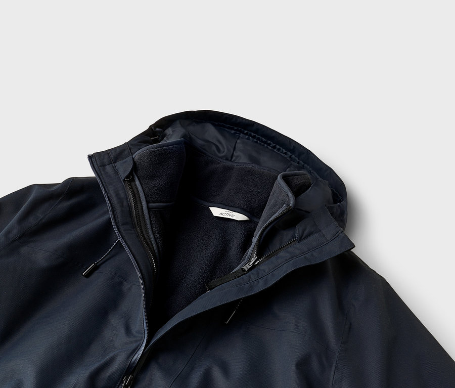 Gros plan d'une veste superposée bleu foncé avec une coque extérieure et une doublure polaire intérieure. La veste est dotée d'une capuche, de plusieurs fermetures éclair et d'une étiquette blanche "ACTIVE" sur la polaire.