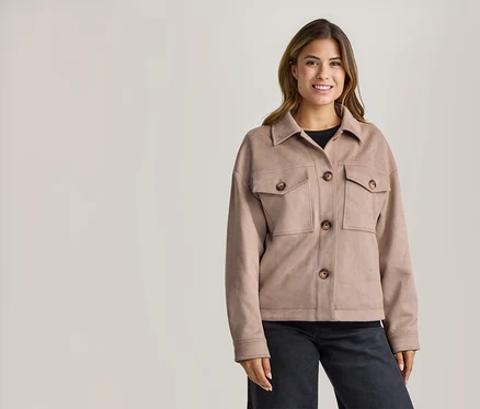 Femme souriante portant une veste marron clair avec un col, quatre boutons marron et deux poches poitrine, un haut noir et un pantalon foncé.