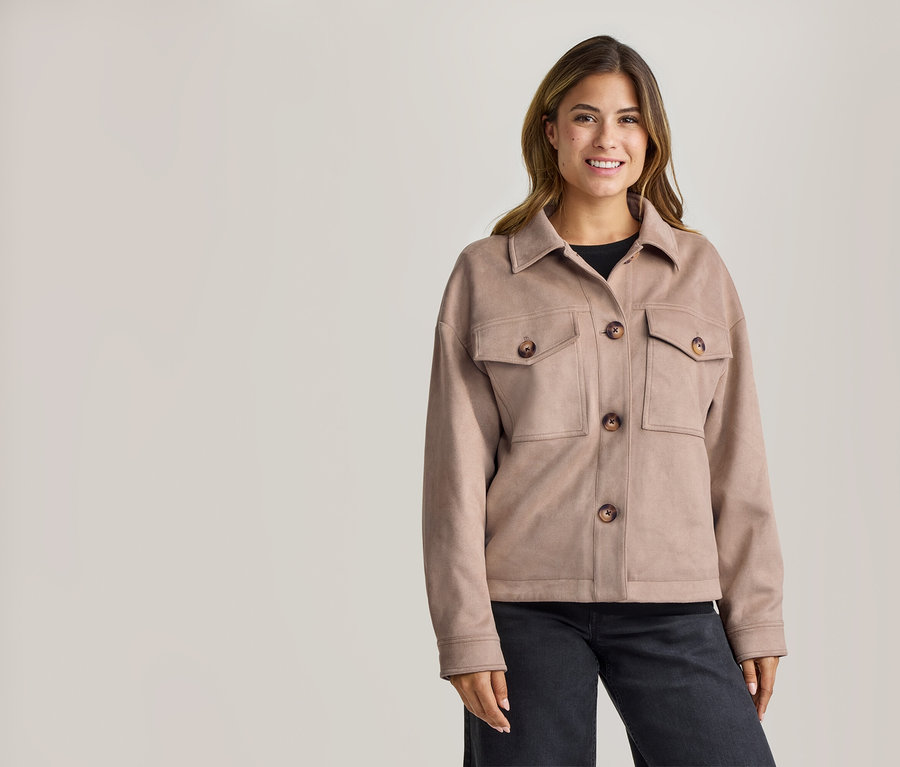 Femme souriante portant une veste marron clair avec un col, quatre boutons marron et deux poches poitrine, un haut noir et un pantalon foncé.