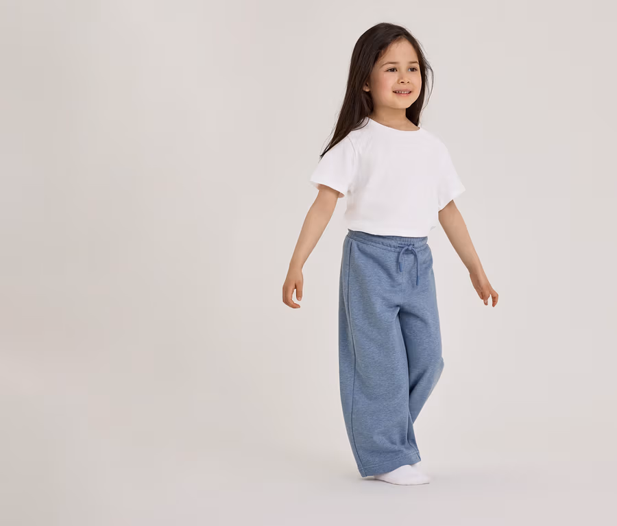 Une jeune fille aux longs cheveux foncés se tient debout, tournée vers la droite, vêtue d'un t-shirt blanc à manches courtes, d'un pantalon de survêtement bleu ample avec un cordon de serrage et de chaussettes blanches.