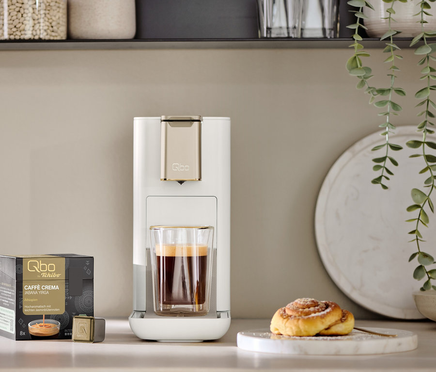 Une machine à café Qbo ICONIC Light Gold blanche avec une tasse de café en dessous, posée sur un comptoir de cuisine. À côté se trouvent un paquet de capsules Qbo Caffè Crema Jabana Yirga et un roulé à la cannelle sur une assiette en marbre.