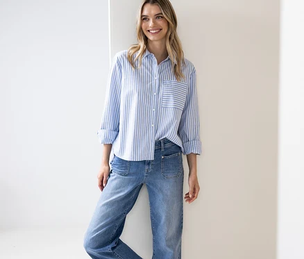 Une femme souriante aux longs cheveux blonds porte un chemisier Street One rayé bleu et blanc avec les manches retroussées et un jean, appuyée contre un mur blanc.