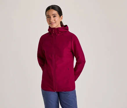 Femme souriante portant une veste à capuche bordeaux et un pantalon bleu, les mains derrière le dos.