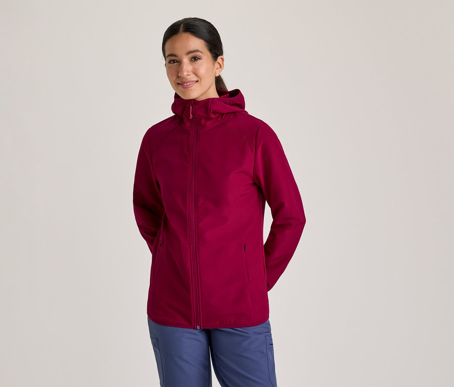 Femme souriante portant une veste à capuche bordeaux et un pantalon bleu, les mains derrière le dos.