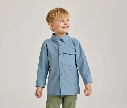 Un jeune garçon aux cheveux blonds sourit et regarde vers la droite. Il porte une chemise bleu clair à manches longues avec une poche et un pantalon vert olive.