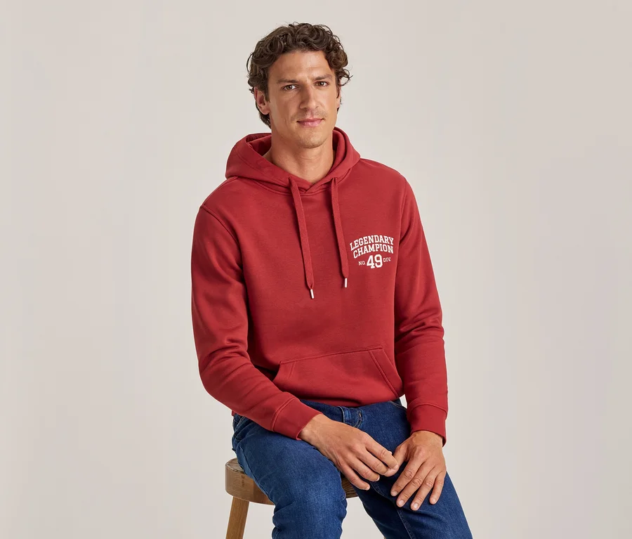 Un homme aux cheveux bouclés est assis sur un tabouret, portant un sweat à capuche rouge avec le texte « LEGENDARY CHAMPION No. 49 DIV » et un jean bleu.