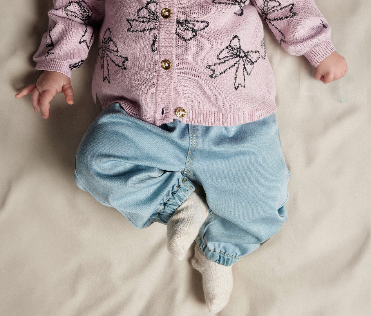 Bébé allongé sur le dos, portant un cardigan rose en tricot avec des motifs de nœuds noirs et des boutons dorés, un pantalon bleu clair et des chaussettes blanches.