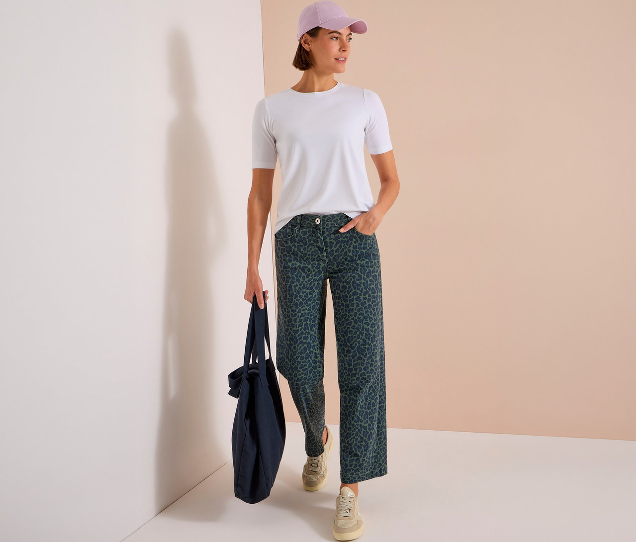 Femme debout, portant un t-shirt blanc, une casquette violet clair et un jean CECIL vert foncé à imprimé léopard, tenant un sac fourre-tout bleu marine.