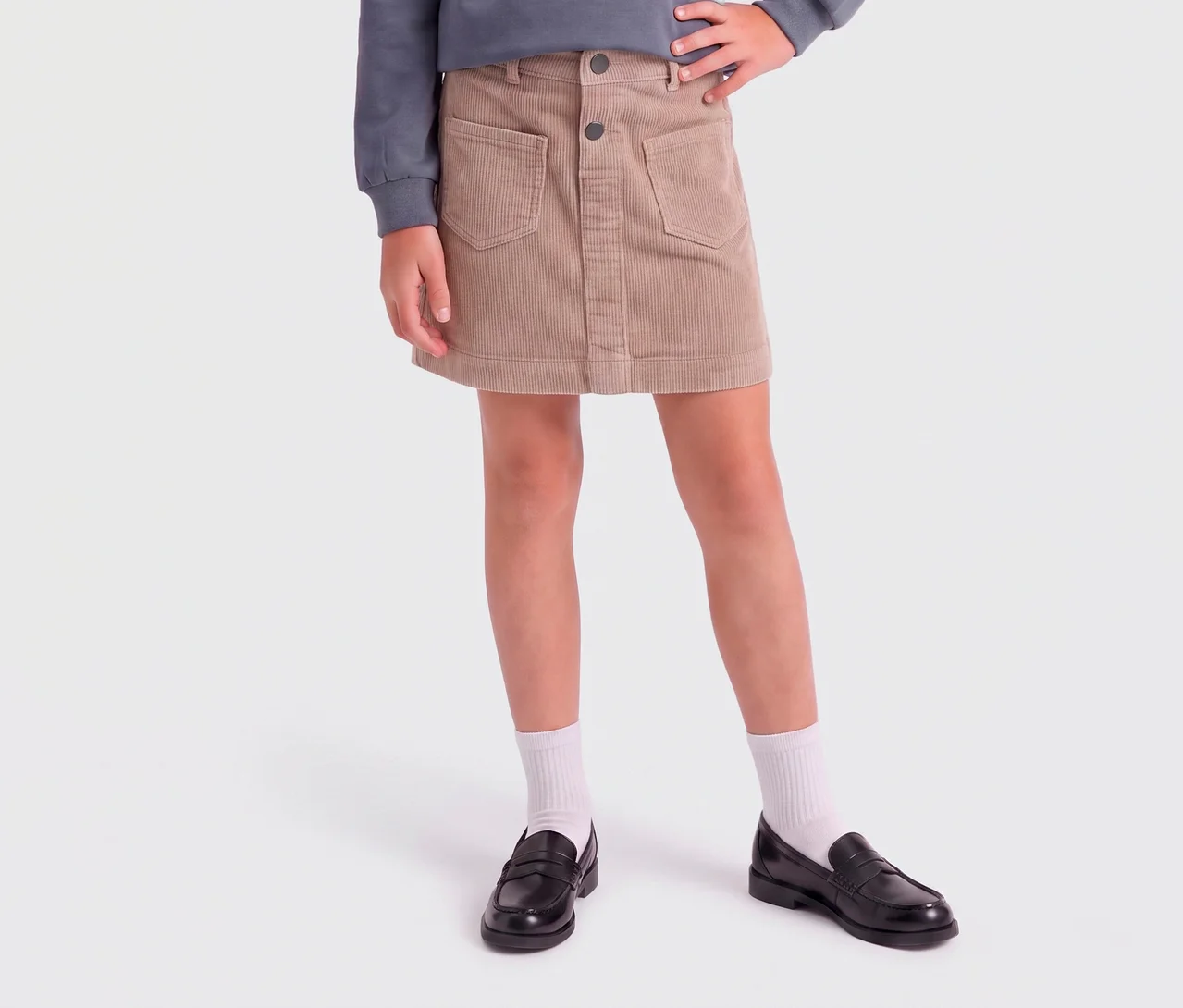 Enfant portant une minijupe en velours côtelé beige avec poches et boutons, un haut gris, des chaussettes blanches et des mocassins noirs.