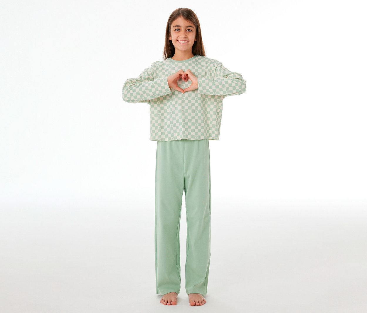 Jeune fille souriante portant un pyjama Interlock pour enfant SCHIESSER, formant un cœur avec ses mains. Elle porte un haut à manches longues à carreaux vert clair et blanc et un pantalon long uni vert clair.