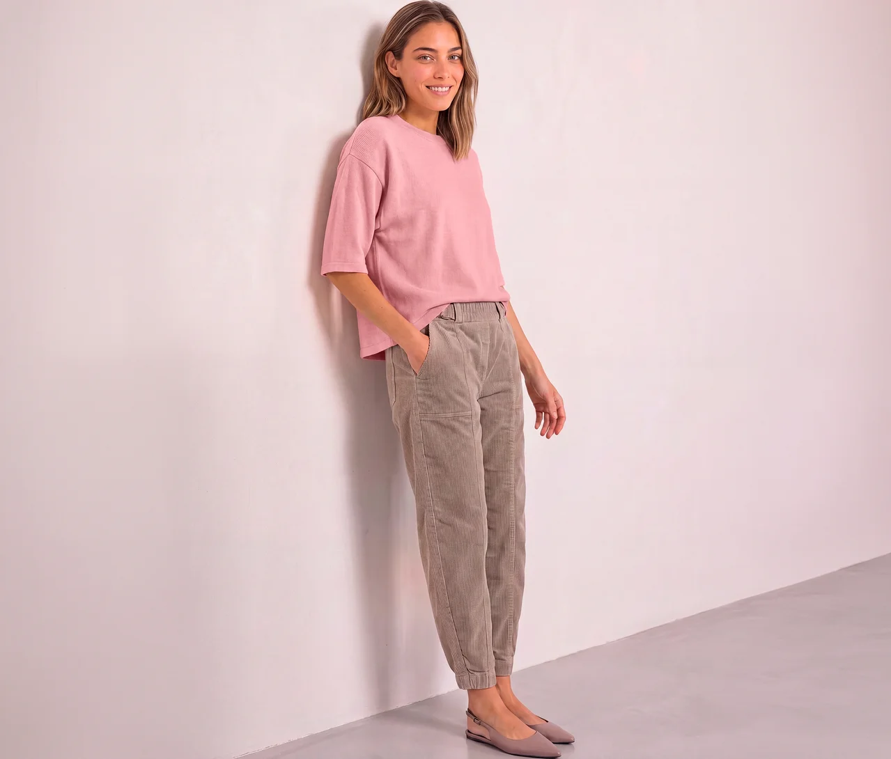 Femme souriante appuyée contre un mur rose, portant un pull en maille 3/4 Street One rose, un pantalon en velours côtelé marron clair et des chaussures plates rose clair.