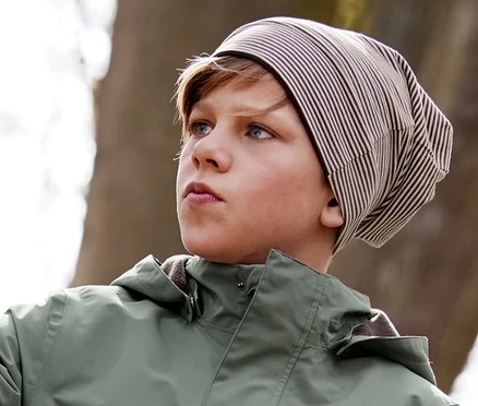 Garçon appuyé contre un arbre avec un bonnet réversible marron, une veste pour tous les temps, un pantalon softshell et des bottes de pluie recyclables marron.