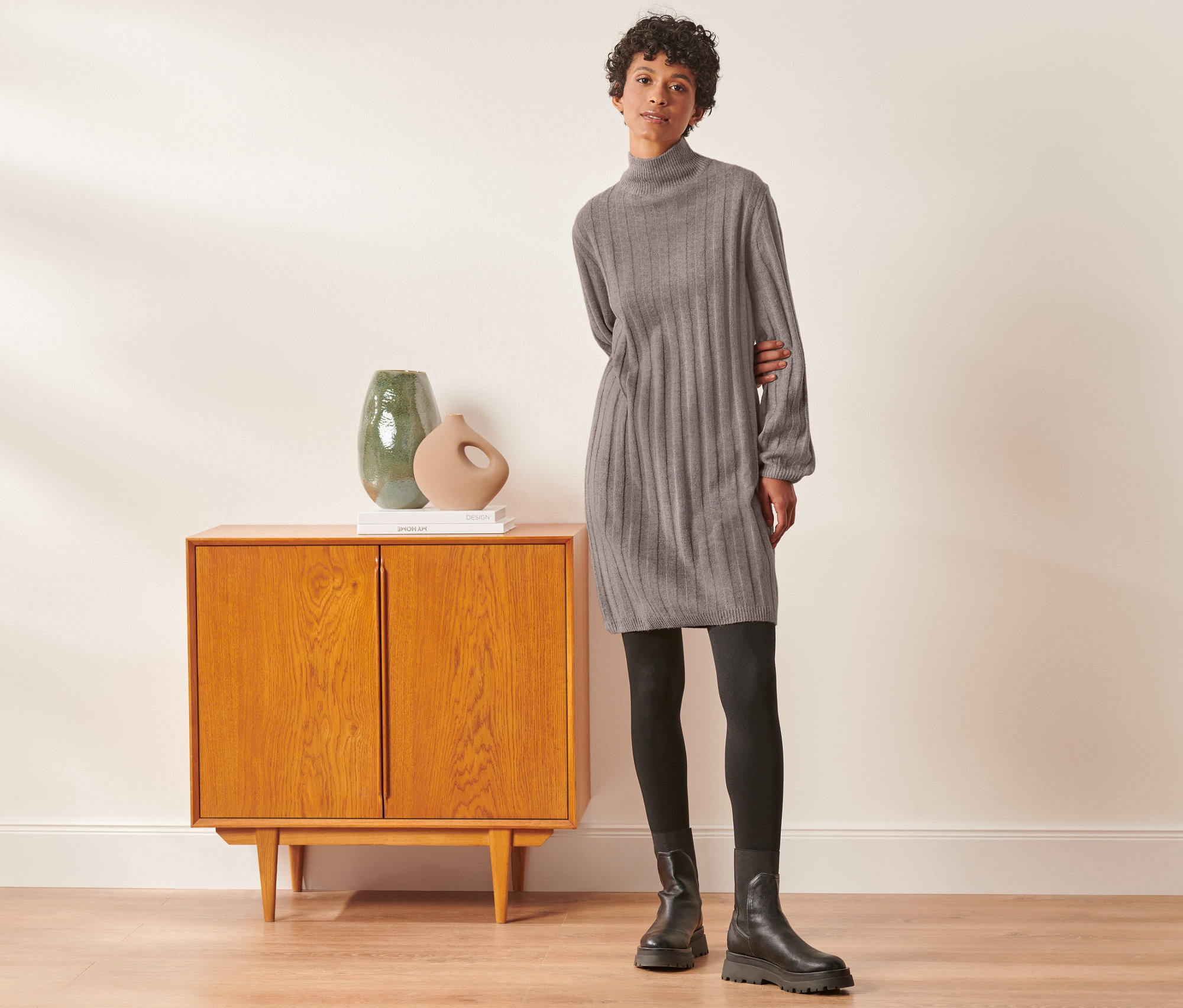 Une femme pose à côté d'une armoire et porte une robe en maille côtelée avec col montant, des leggings noirs et des bottes.