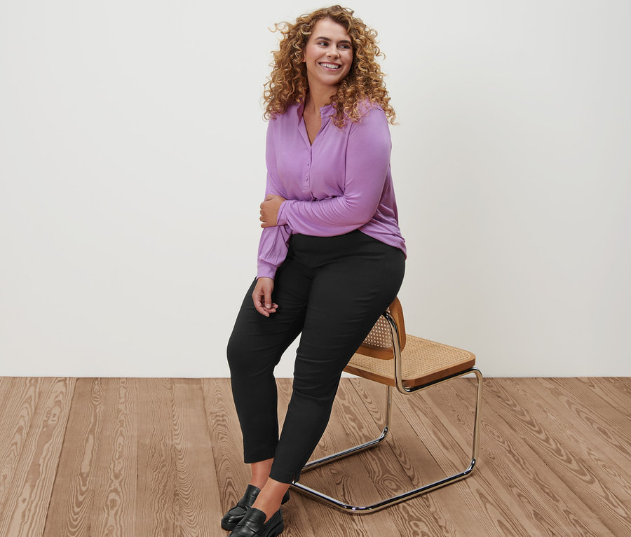 Une femme est assise sur une chaise et porte une blouse lilas avec patte de boutonnage, un pantalon stretch noir et des loafers en cuir.