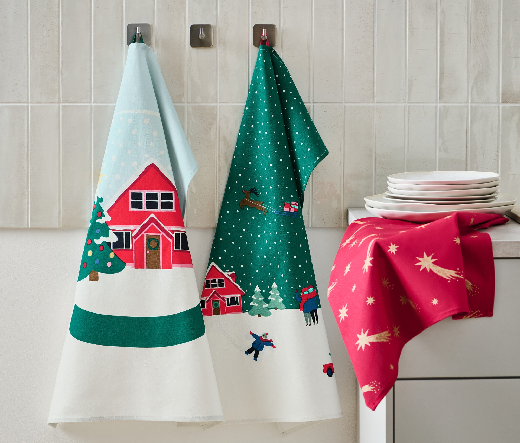 Trois linges de cuisine avec des motifs de Noël sont accrochés au mur. Des assiettes et un distributeur de savon sont posés sur le plan de travail.