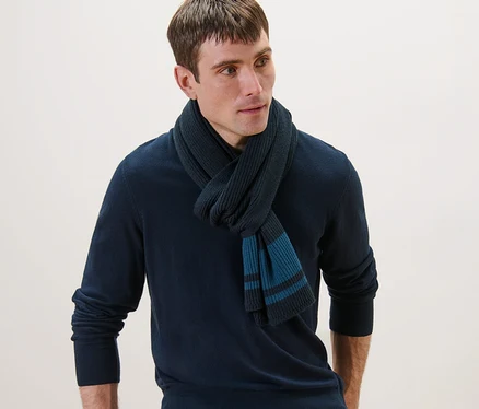 Un homme portant une écharpe en maille, un pull bleu marine et un jean, appuyé sur une chaise.
