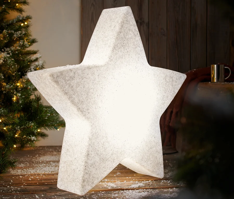 Une lampe smart d’extérieur « étoile », L, illuminée est posée sur un plancher en bois enneigé à côté d’un sapin de Noël.