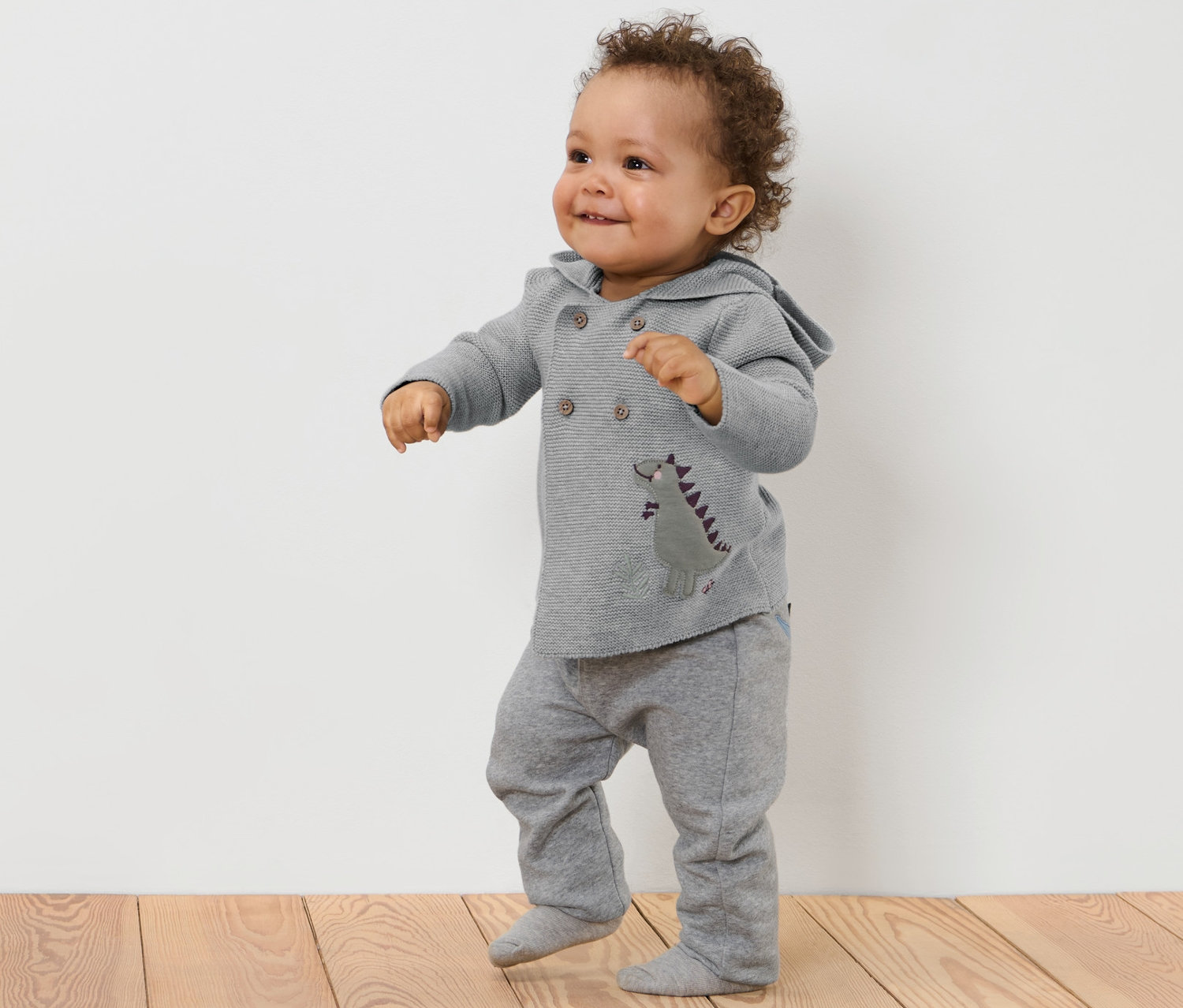 Un bébé souriant portant une jaquette en maille pour bébé avec motif appliqué et un pantalon de jogging gris se tient sur un plancher en bois.