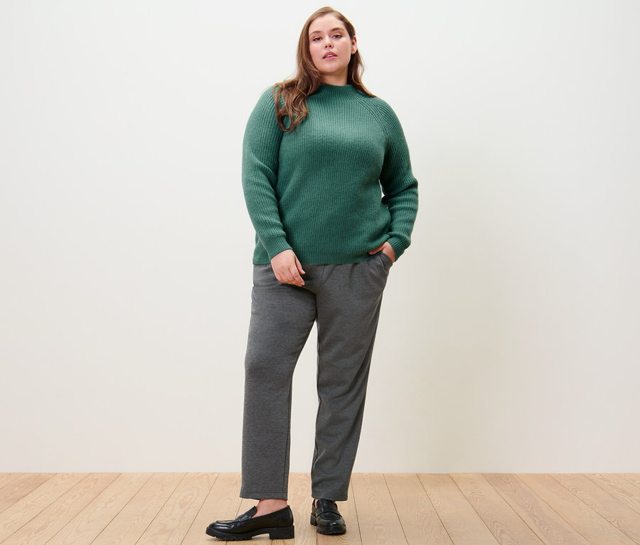 Femme posant en pull à col droit en grosse maille vert et pantalon de jogging.