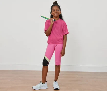 Une fille avec une raquette de badminton sur l'épaule porte 2 t-shirts de sport enfant, des leggings roses et des baskets blanches.