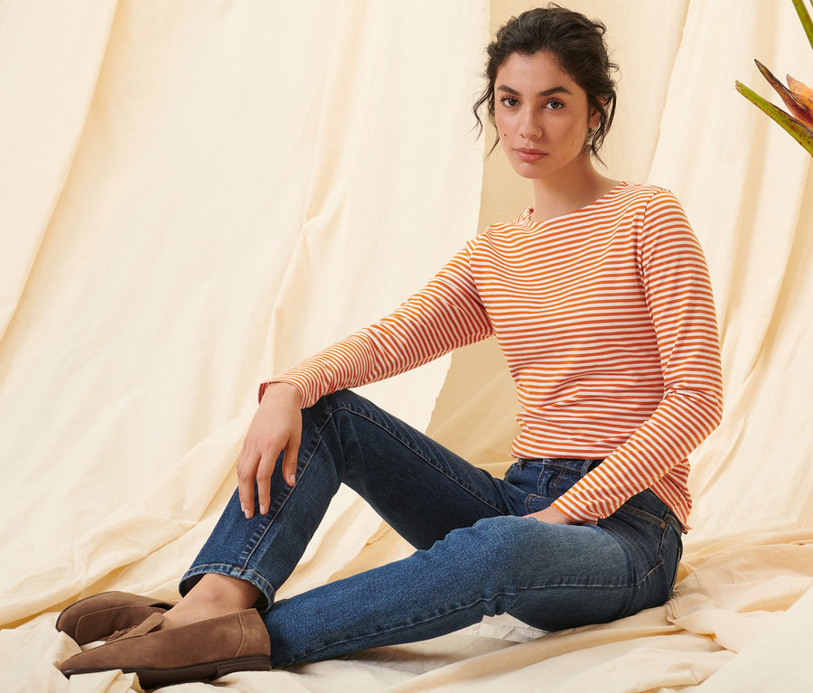 Femme assise portant un t-shirt manches longues à rayures orange, un jean droit en denim et des loafers en cuir velours.