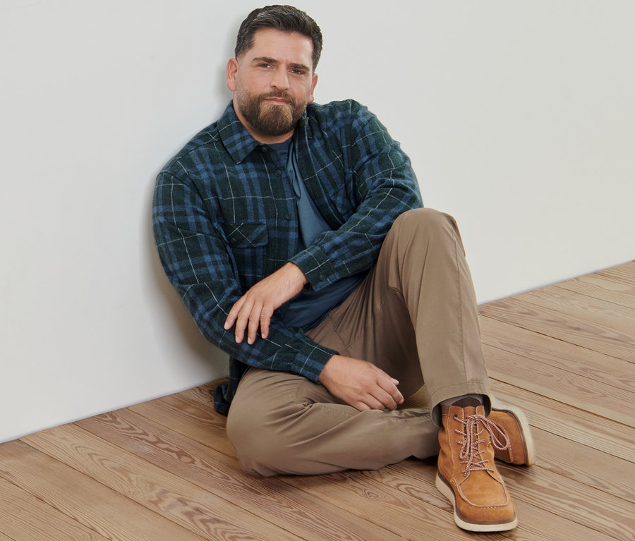 Un homme est assis sur un plancher en bois et porte un pantalon chino de couleur sable, une veste-chemise en flanelle et 2 t-shirts manches longues.