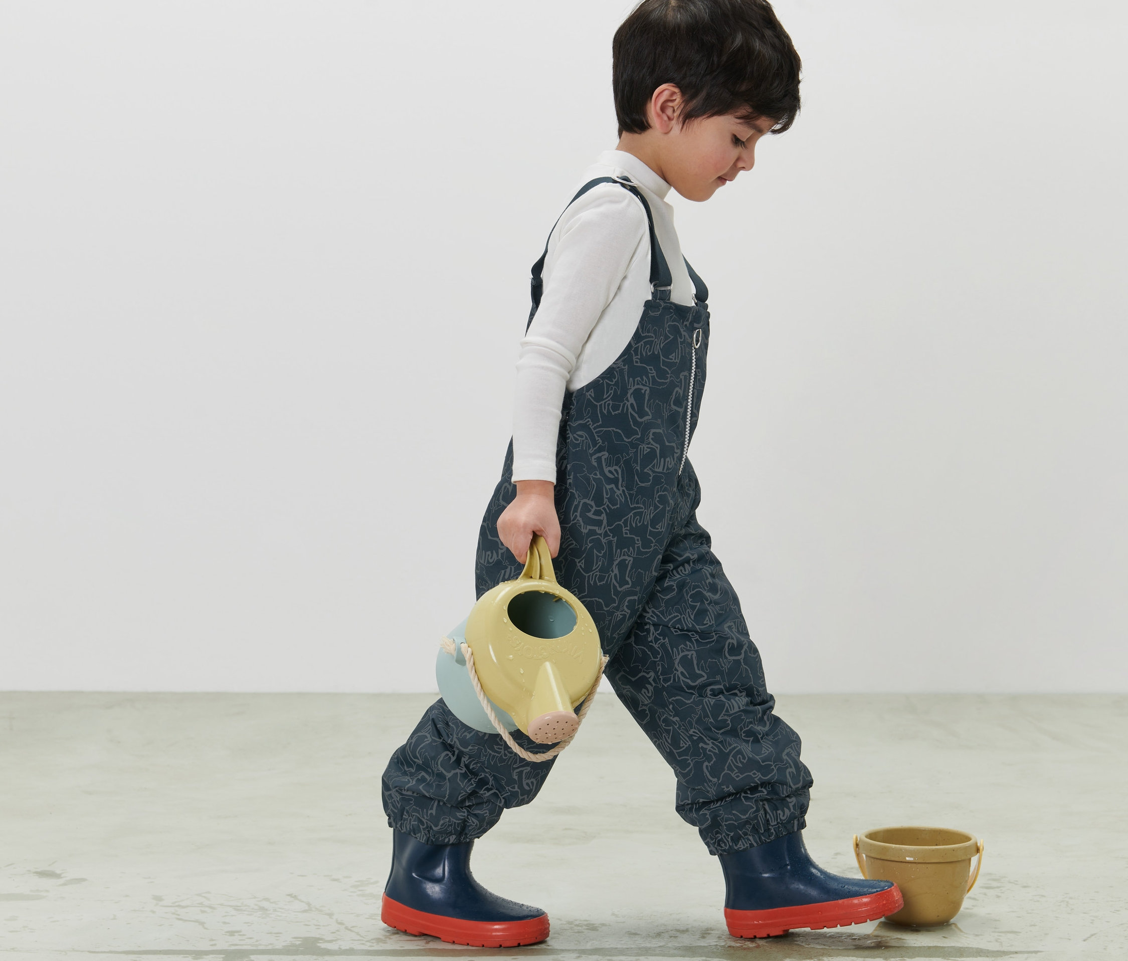 Un garçon marche en portant un arrosoir. Il porte un pantalon de pluie thermique bleu foncé pour enfant avec des éléments réfléchissants.