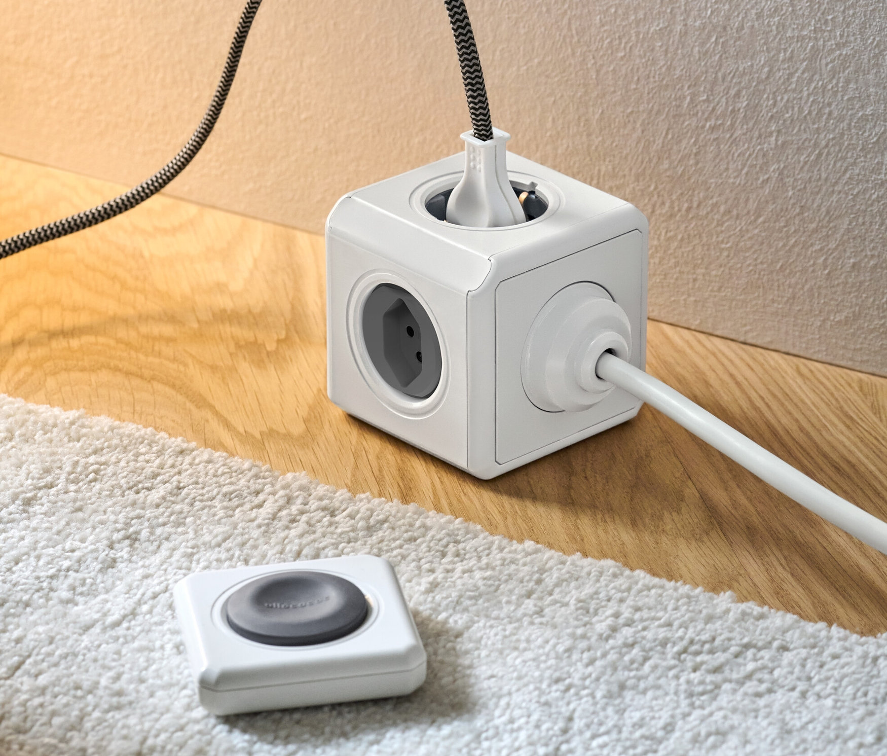 Un cube multiprise blanc avec télécommande et des câbles branchés sur un plancher en bois et un tapis blanc.