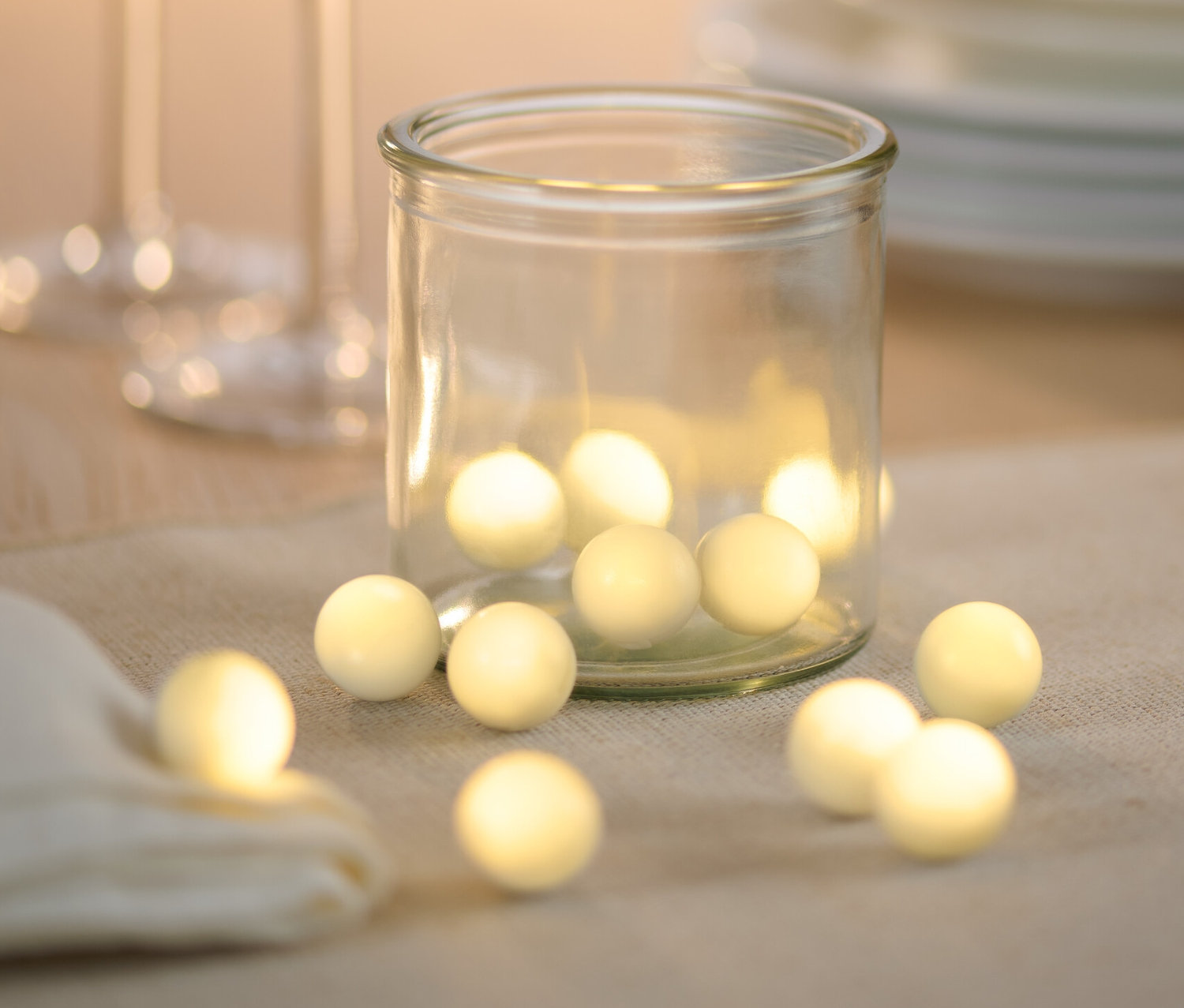 Un verre avec 12 petites boules à LED sur une table à côté de verres et d'assiettes.