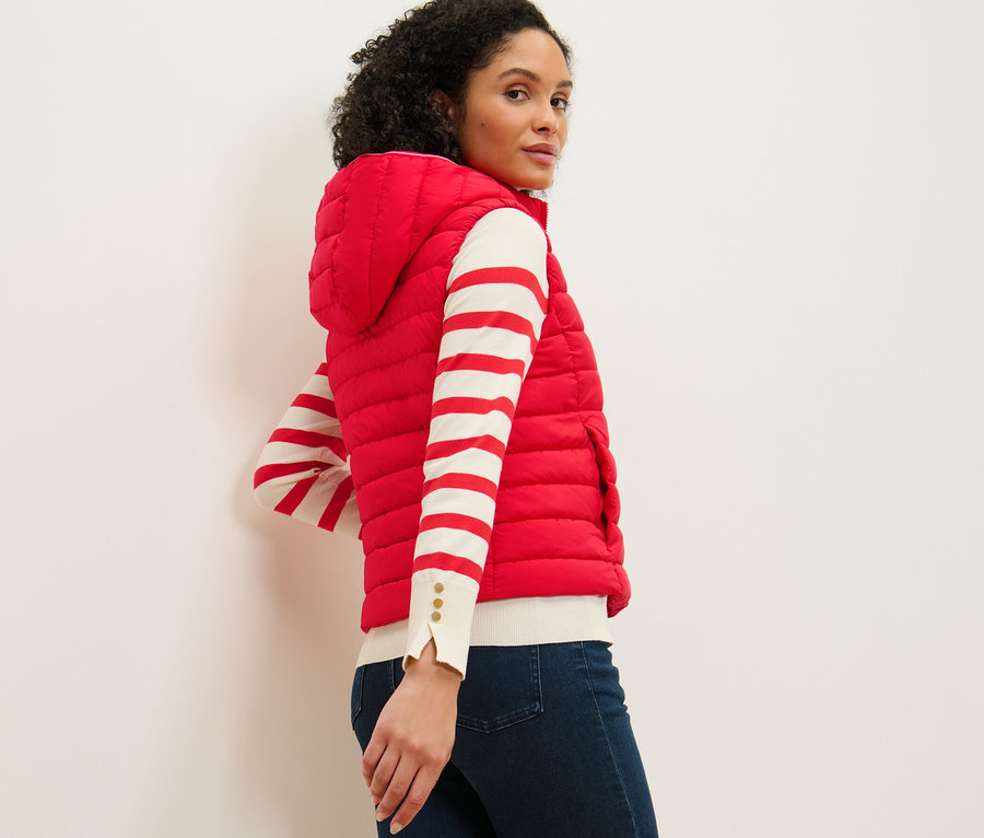 Une femme regarde par-dessus son épaule et porte un gilet sans manches matelassé à capuche, un haut rayé et un jean.