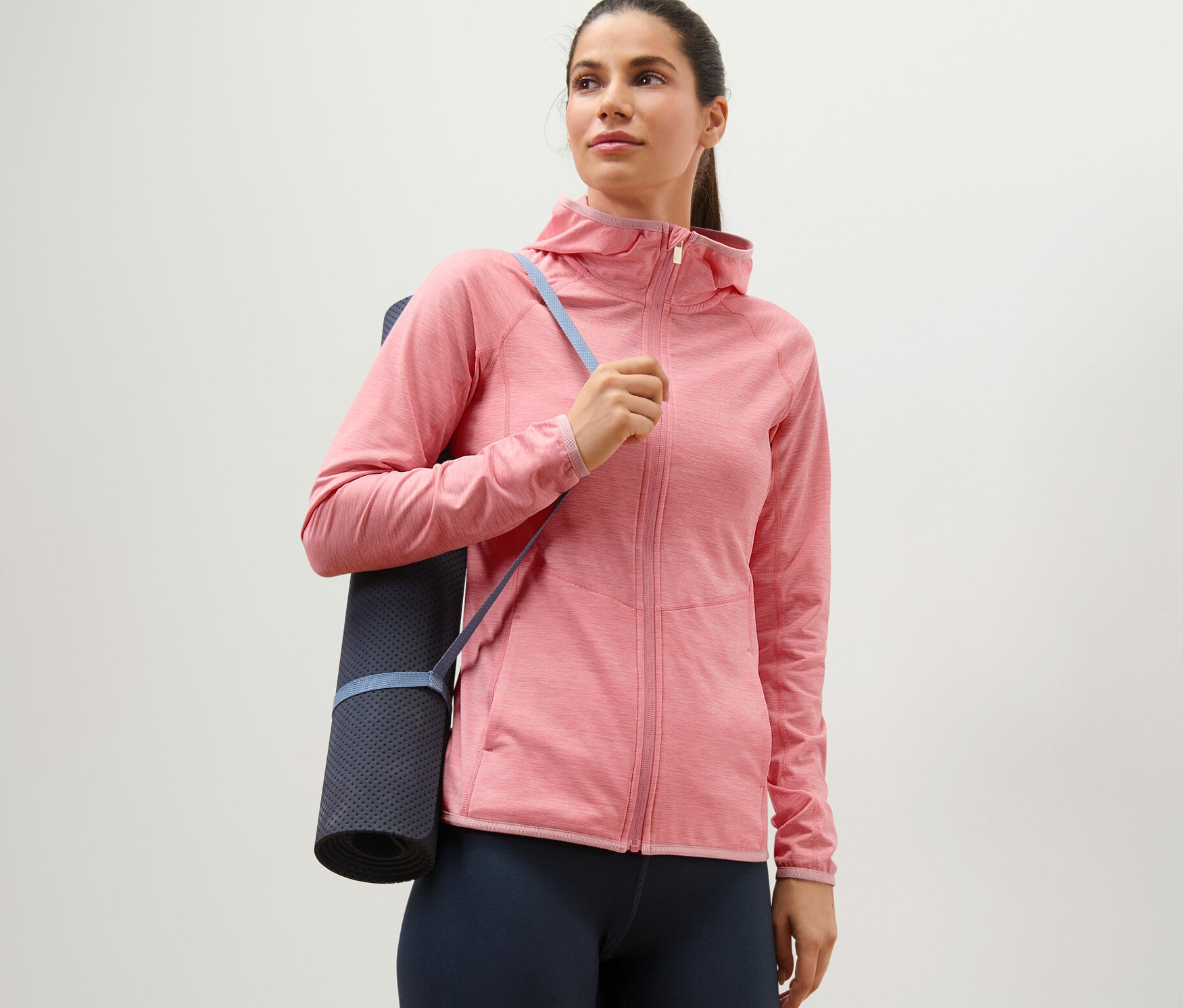 Une femme portant une veste de sport rose chiné et un tapis de yoga sur l'épaule.