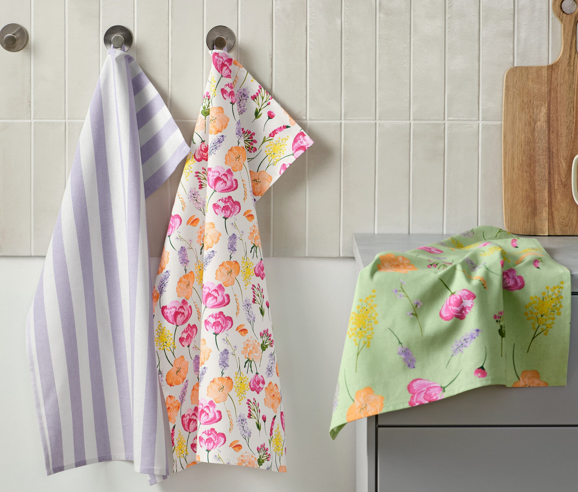 Trois torchons sont accrochés au mur : un torchon rayé violet et blanc, un avec un motif floral et un sur le comptoir. À côté se trouvent une planche à découper et une tasse avec des ustensiles de cuisine. 3 linges de cuisine