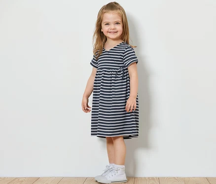 Une fille souriante se tient de côté et porte une robe en jersey pour enfant avec des rayures bleues et blanches et des baskets blanches.