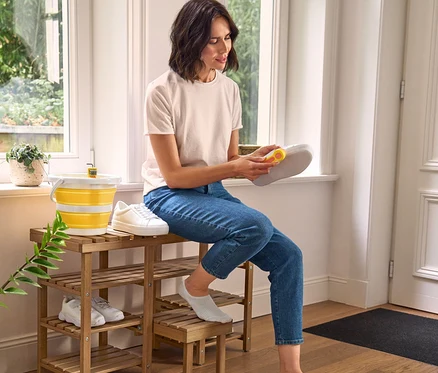 Une femme nettoie une chaussure avec une brosse à chaussures 3 en 1, assise sur un banc à chaussures haut avec marchepied rabattable. À côté d'elle se trouvent une brosse à chaussures et un seau pliable.