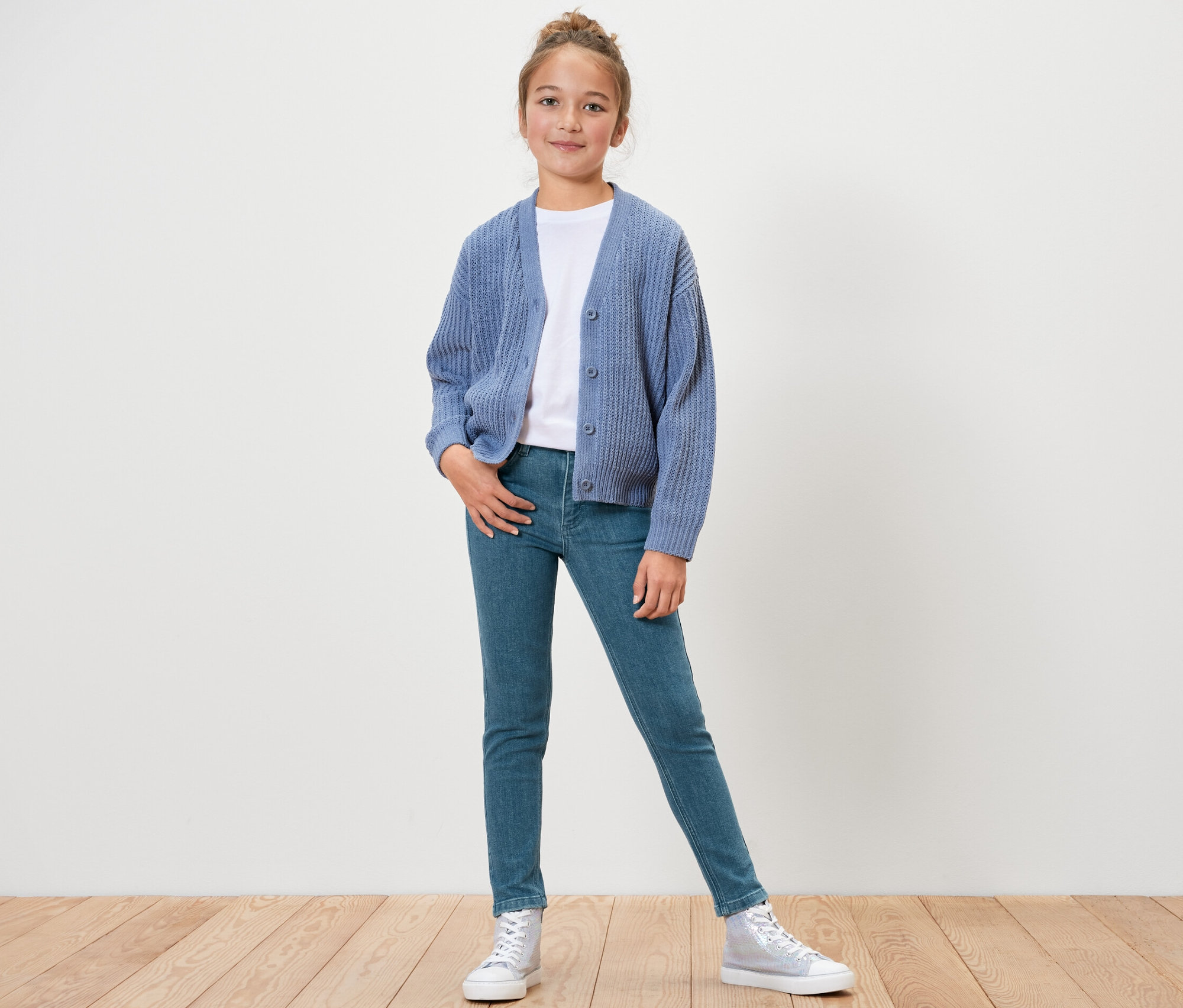 Fillette posant debout en jean pour enfant – Fit « Lotta », t-shirt blanc et cardigan bleu.